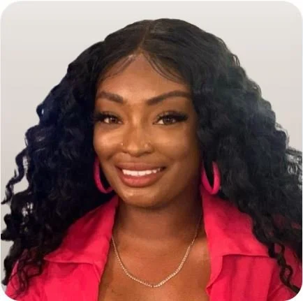 A woman with long curly black hair, wearing pink hoop earrings, a pink top, and a silver necklace, smiling at the camera.