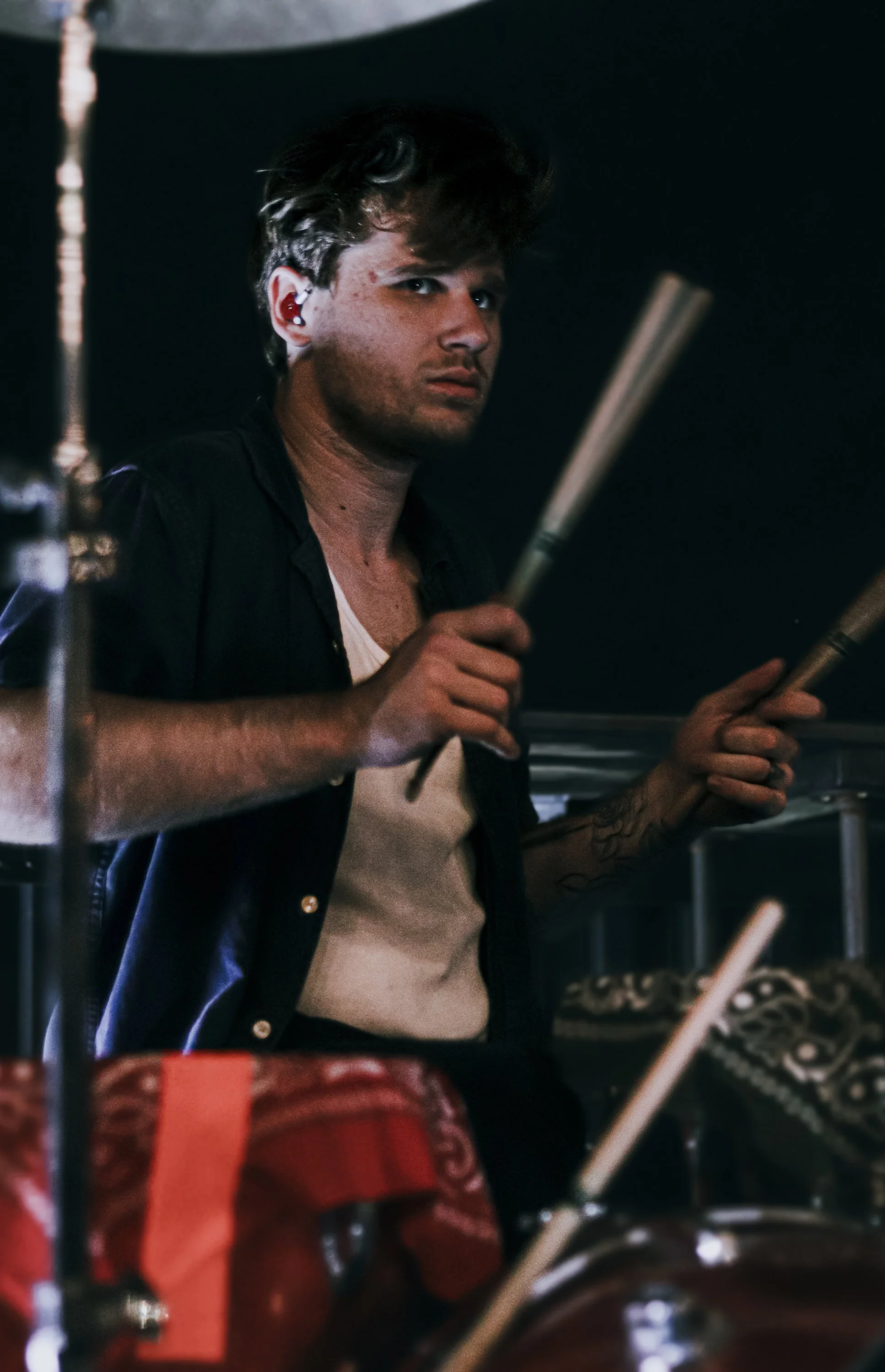 A young man with wavy hair holding drumsticks, looking towards the camera in a dark setting.