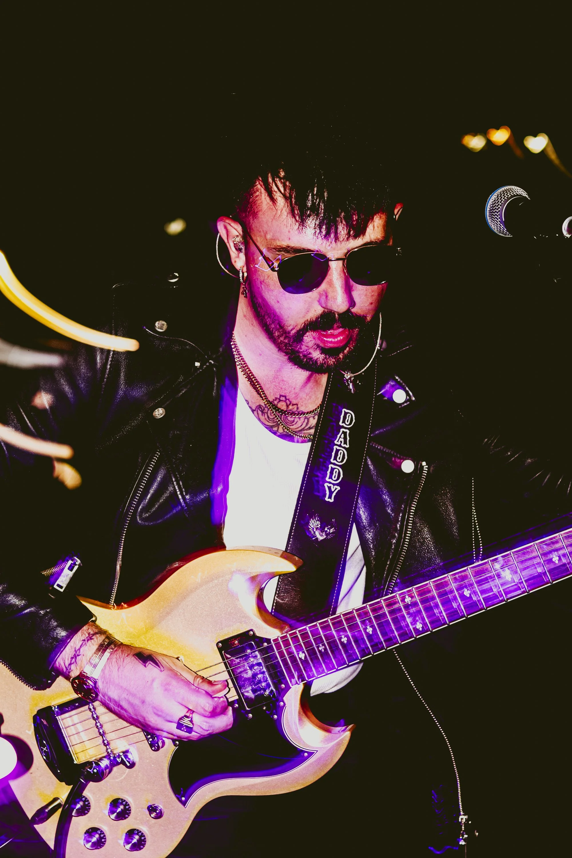 A male guitarist with dark hair and sunglasses playing an electric guitar on stage, wearing a leather jacket and a strap with the word 'DADDY' on it.