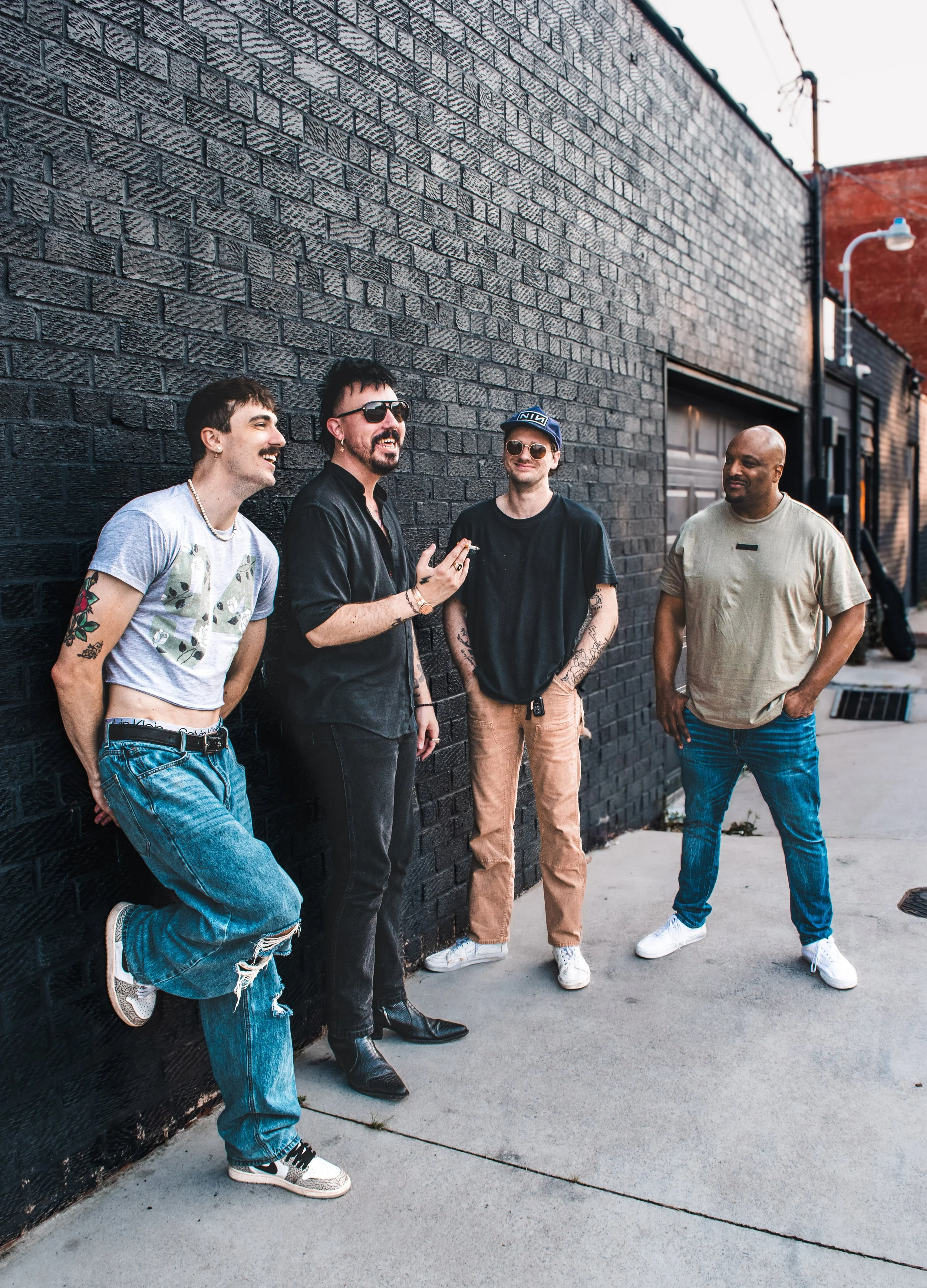 Four men stand talking and laughing outside against a black brick wall, dressed casually with tattoos and sunglasses, on a city sidewalk at sunset.