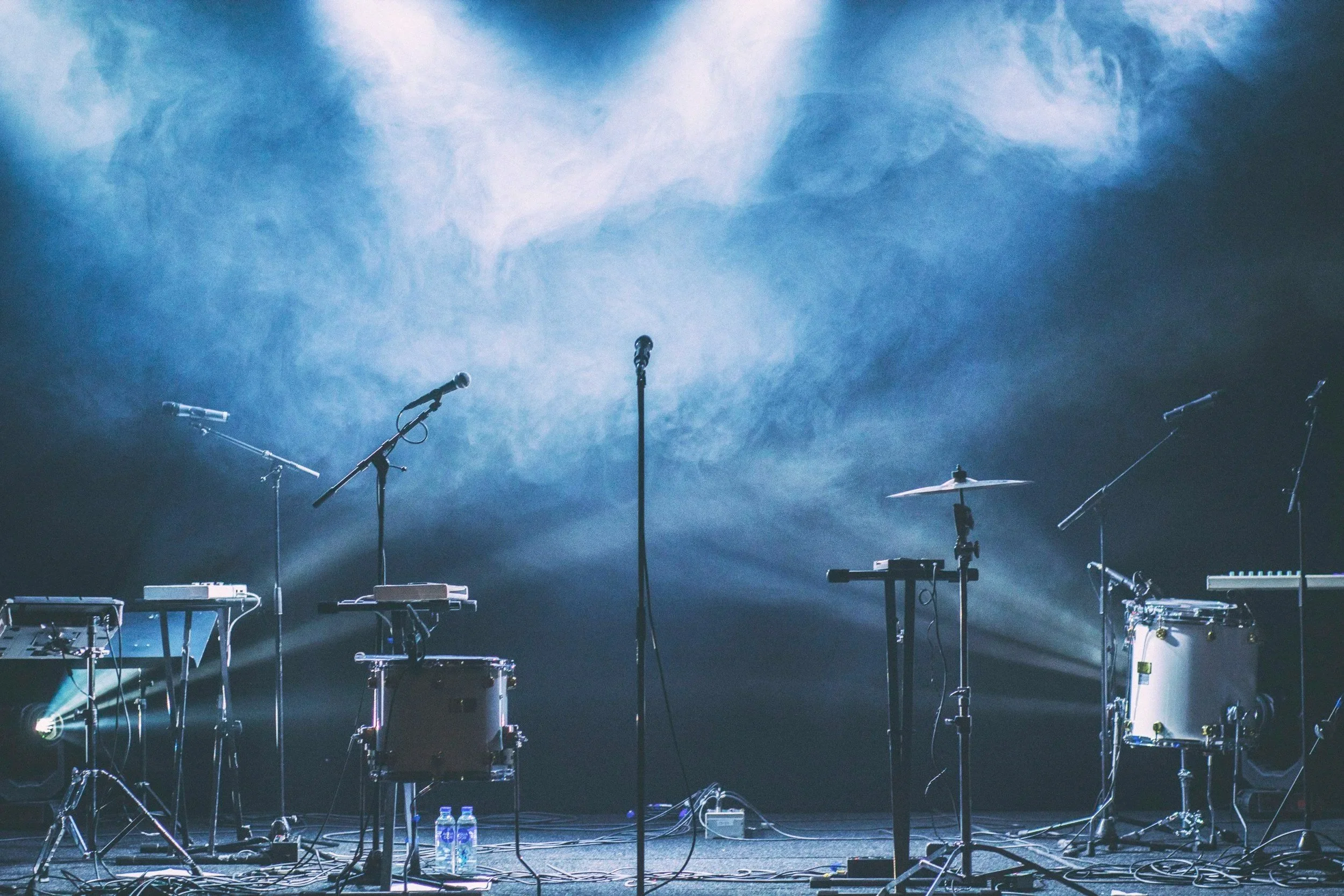 Empty stage with musical instruments and microphones, illuminated by stage lights, with a blue cloudy background.