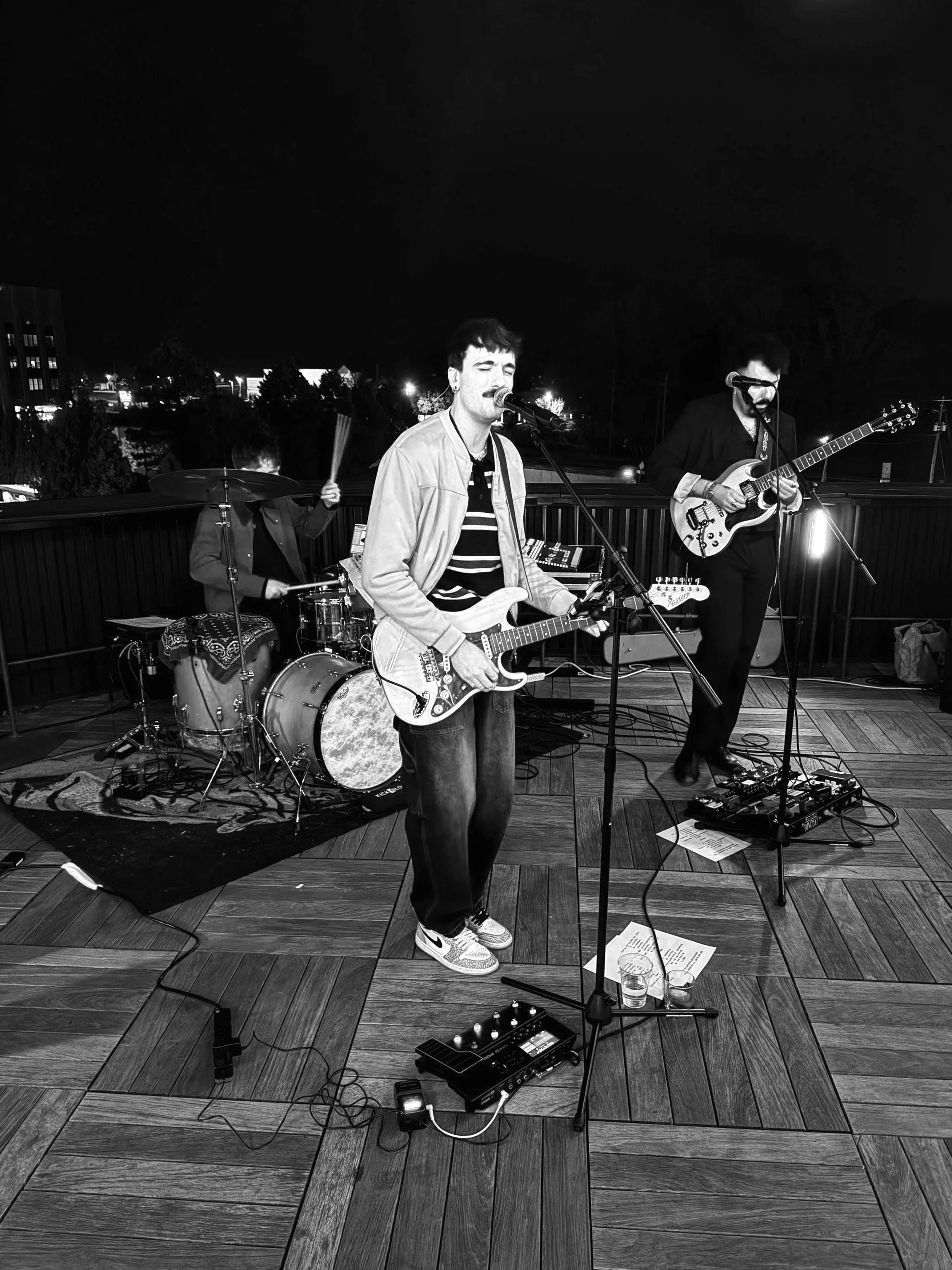 A band performing live on a wooden stage at night, with a guitarist, bassist, and drummer, and various musical equipment and papers on the floor.