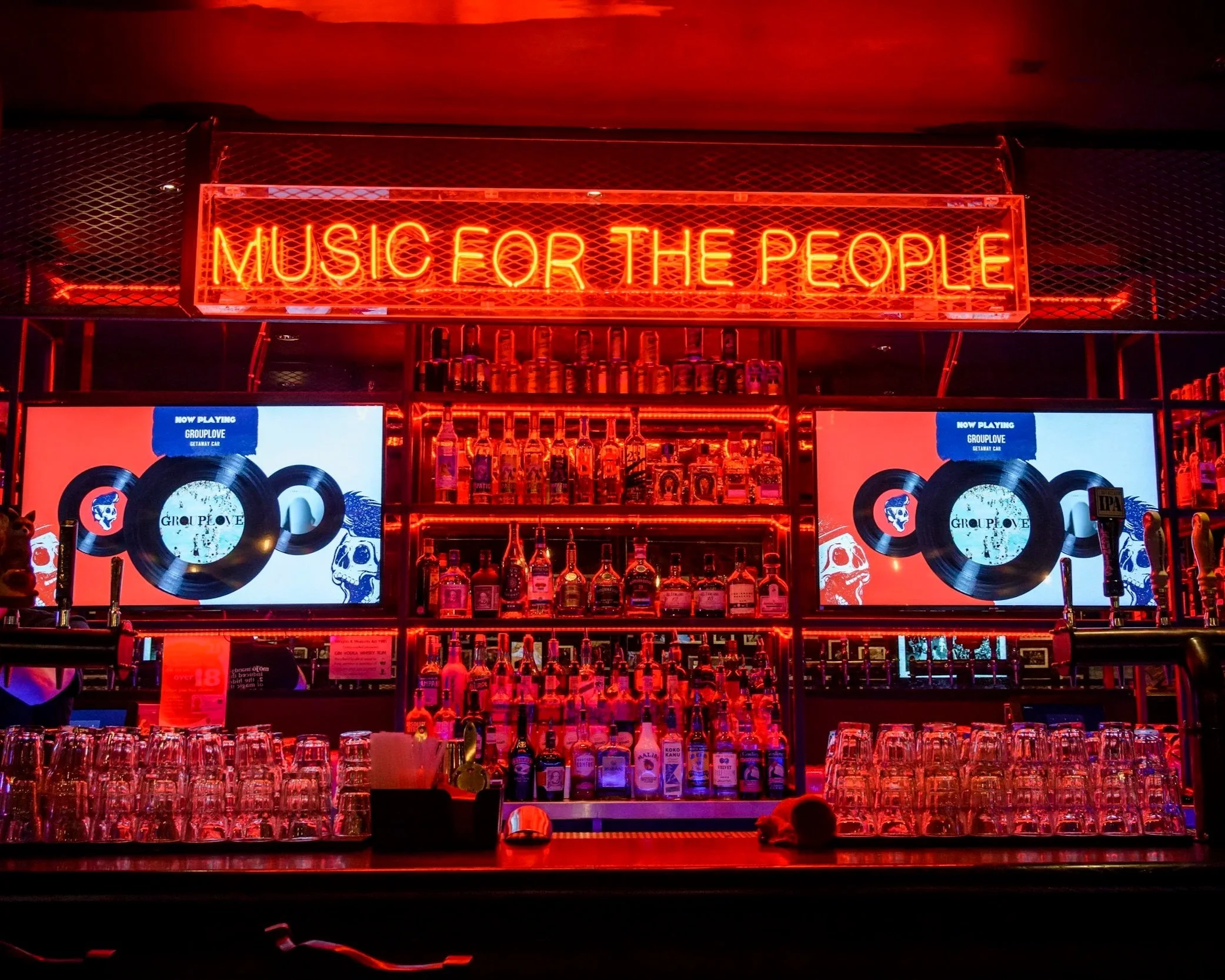 Neon sign reading 'Music for the People' above a bar with bottles of liquor, glasses, and two screens displaying vinyl record graphics and the word 'Grouplove' in a dimly lit, red-hued environment.