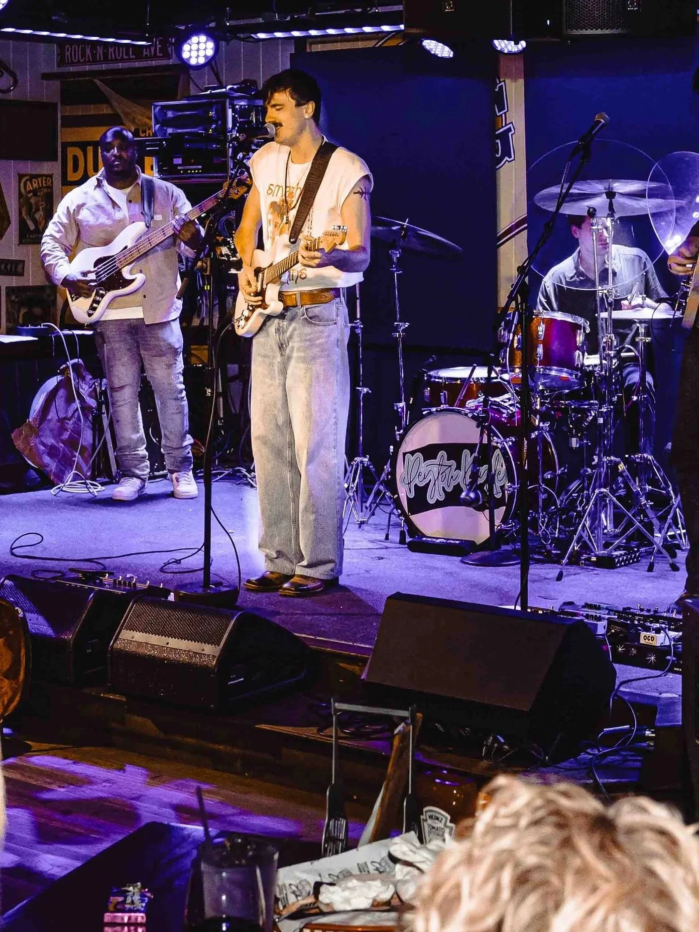 A band performs on stage with a singer-songwriter playing an electric guitar at the center. The band includes a bassist on the left and a drummer behind the singer. The stage has music equipment, microphones, and a backdrop with a black screen.