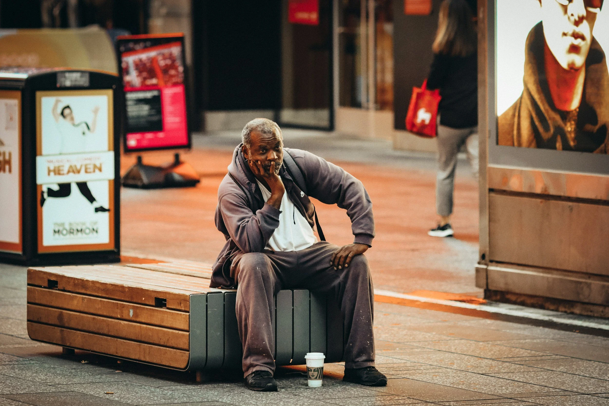 An elderly man sits on a wooden bench at night, with a coffee cup on the ground in front of him, in an urban area, looking contemplative with his hand on his face.