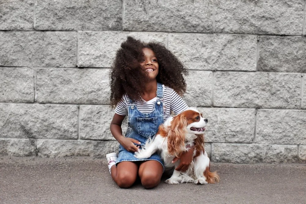 A young girl with curly hair, wearing a striped shirt and denim overalls, sitting on the ground with her hand on a Cavalier King Charles Spaniel dog, in front of a stone wall.