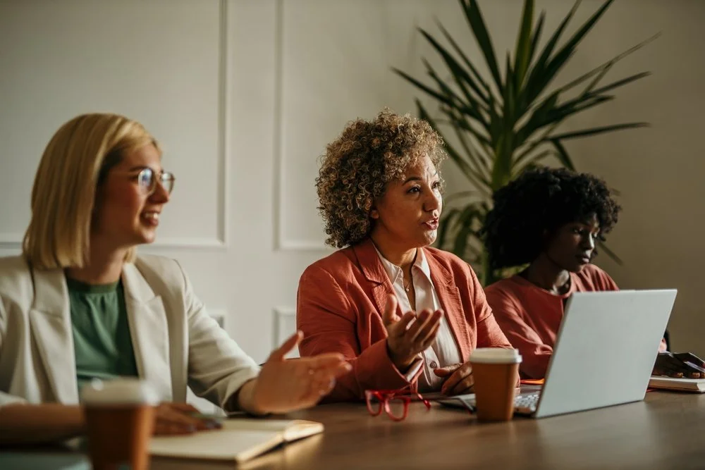 Three women seated at a table in a meeting or discussion, with coffee cups and notebooks, one woman speaking and gesturing.