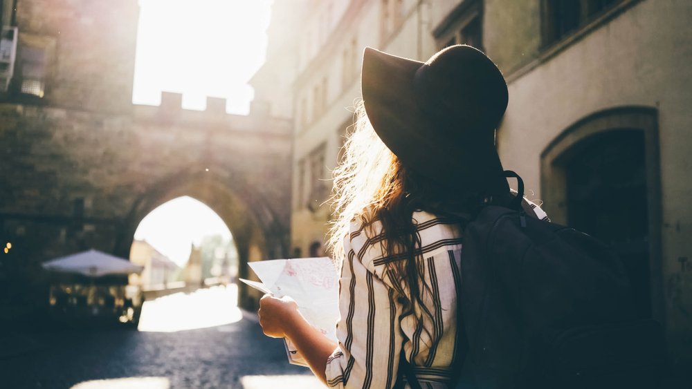 A woman with long hair wearing a wide-brimmed hat and striped shirt, carrying a backpack, looking at a map in an outdoor city setting with sunlight shining through an archway.