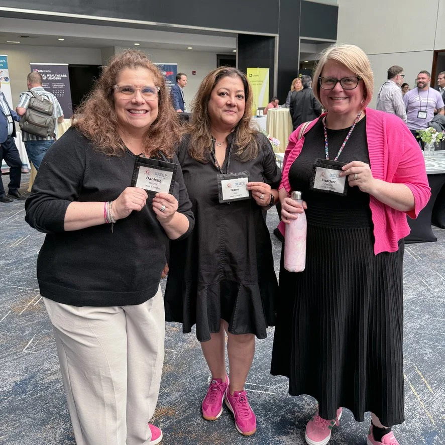 Three women at a conference, wearing name tags and holding small items, standing in a busy indoor event space.