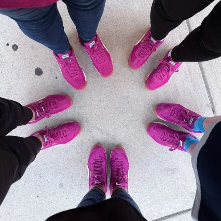 Group of people standing in a circle wearing matching bright pink sneakers.