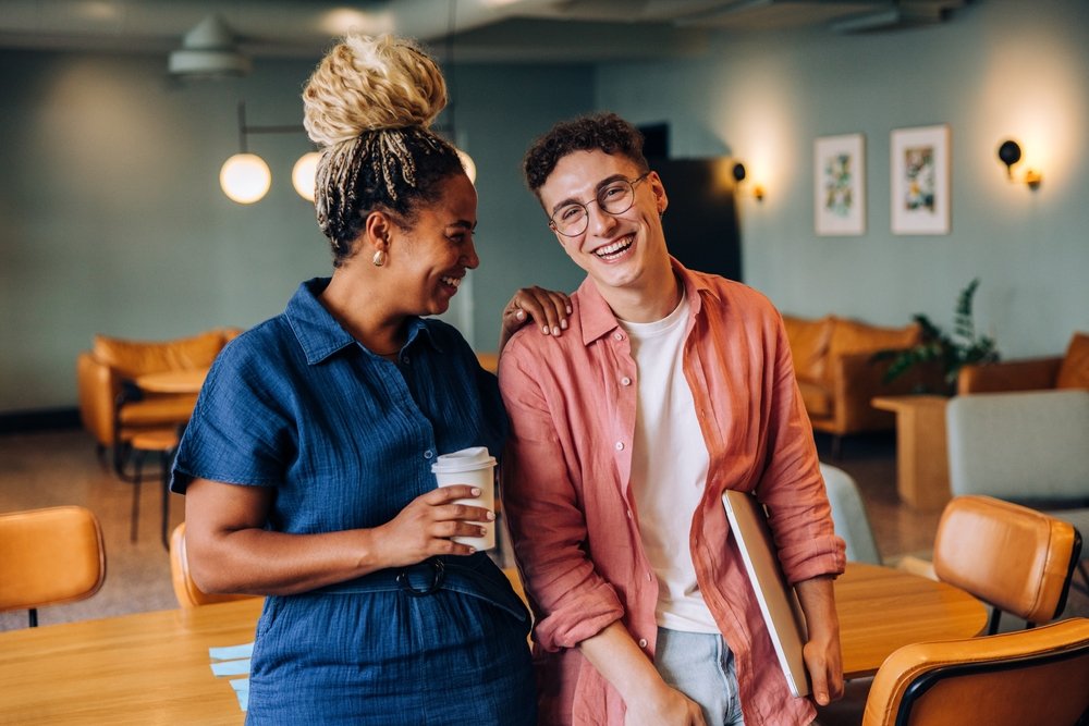 Two people smiling and enjoying conversation in a coffee shop, one holding a cup of coffee and the other carrying a laptop.
