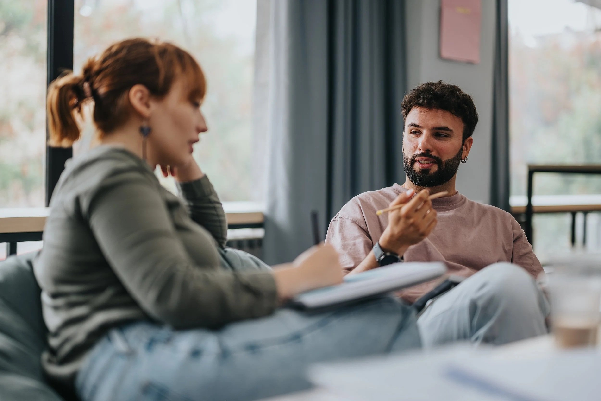A man and a woman are sitting in a room near a large window, engaged in a conversation. The woman, with red hair tied back and wearing a striped shirt, seems to be listening, while the man, with dark hair and beard, is holding a pencil and speaking.