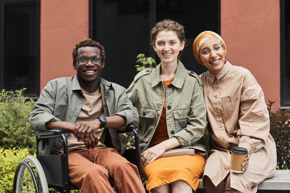 Three diverse friends sitting together outside, smiling, with one using a wheelchair and holding a coffee cup.