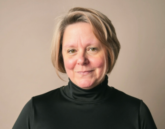 A woman with short blonde hair wearing a black turtleneck sweater standing against a beige background.