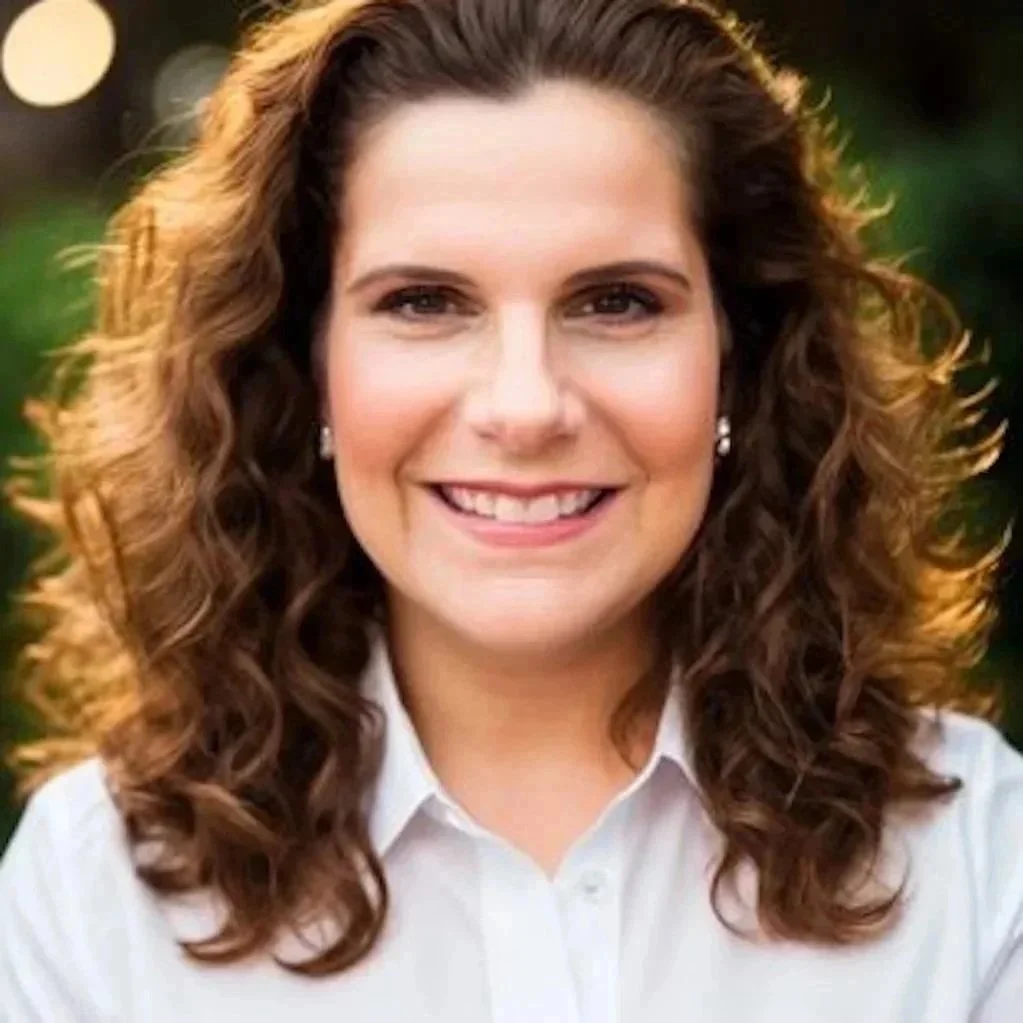 A woman with curly brown hair smiling outdoors, wearing a white collared shirt and pearl earrings.
