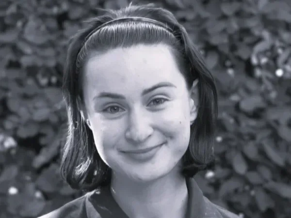 A black and white photo of a smiling young woman with dark hair held back with a headband, standing outdoors with blurred foliage in the background.