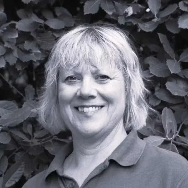A woman with short, wavy, gray hair smiling in front of leafy bushes.
