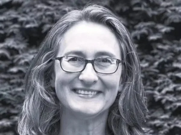 A woman with wavy hair and glasses smiling in front of a leafy background.