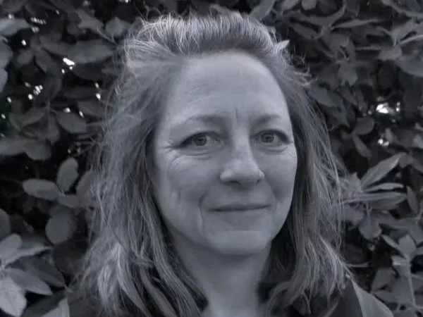 A black and white photo of a woman with shoulder-length hair standing in front of leafy bushes.