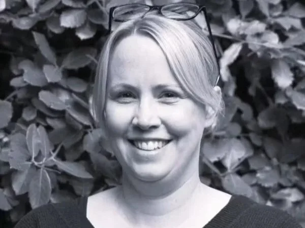 A woman with glasses on her head smiling outdoors in front of leafy foliage.