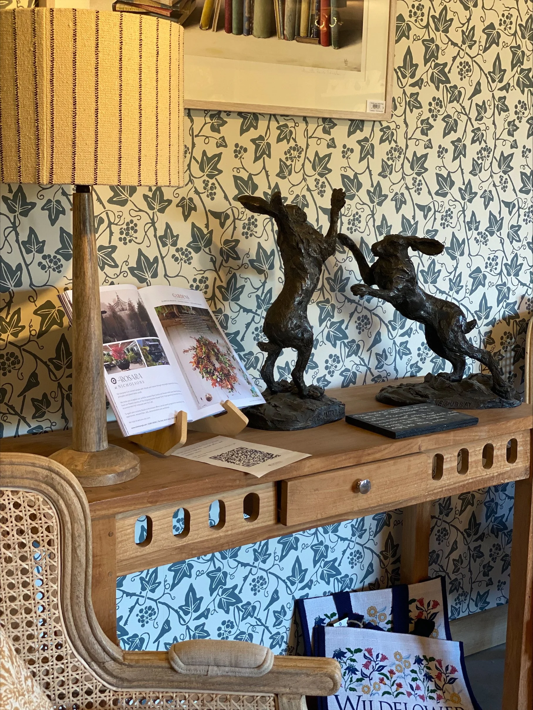 Bronze hare sculptures, a table lamp, an open magazine showcasing floral arrangements. The background features a wallpaper with a dark leaf pattern.