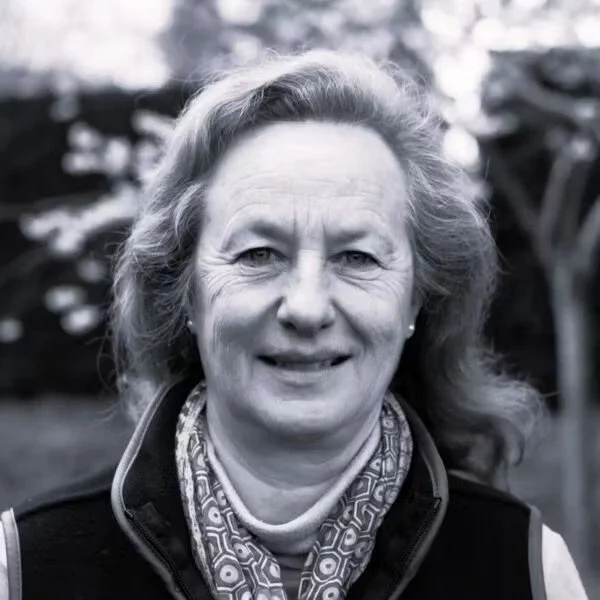 A middle-aged woman with wavy hair smiling outdoors, wearing a patterned scarf and a vest.