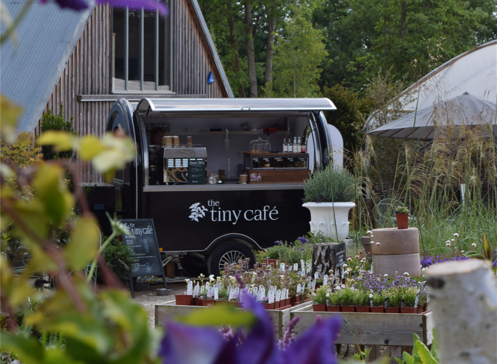 A small black coffee cart labeled 'the tiny café' is set up outdoors among flowers and plants, with a wooden building and trees in the background.