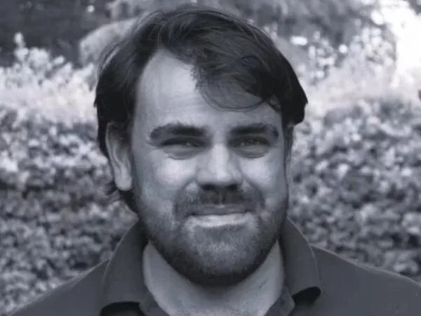 A black-and-white photo of a man with dark hair and a beard, smiling, outdoors with a blurred background of trees and foliage.