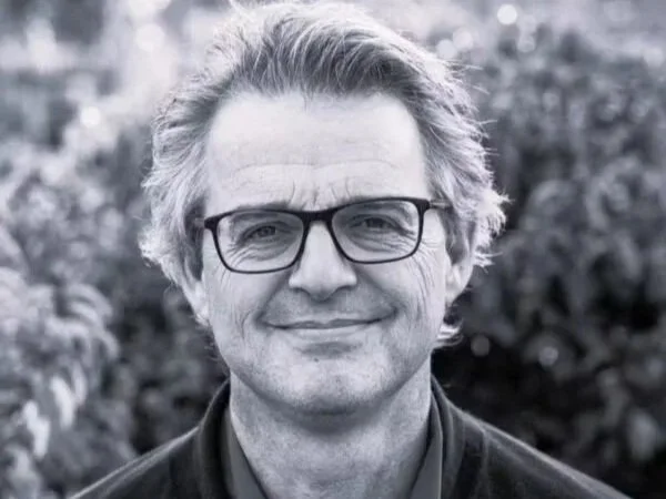 A black-and-white portrait of a middle-aged man with glasses, smiling, outdoors with blurred foliage in the background.