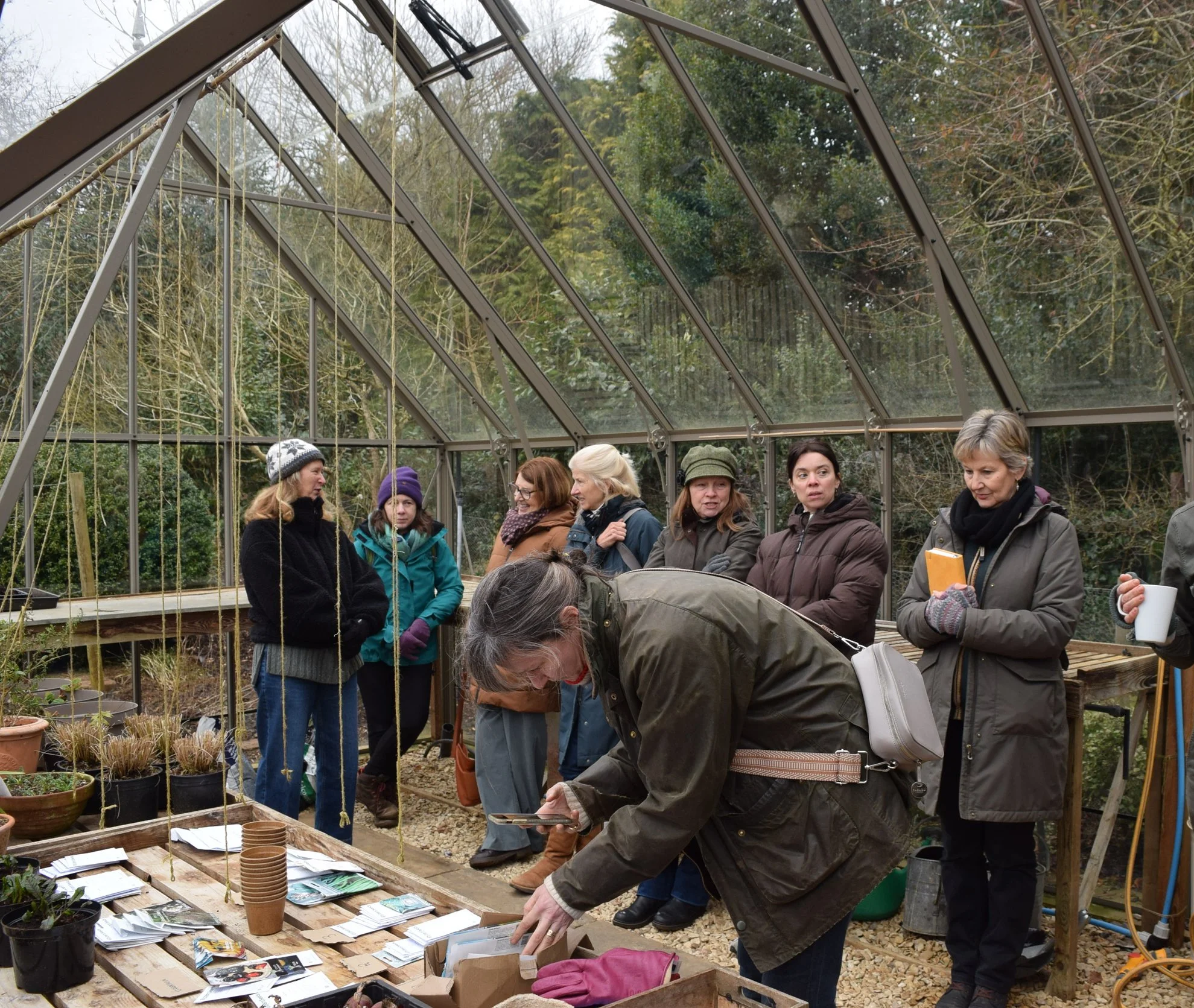 Kathy Slack workshop - Group of people gathering inside a greenhouse, looking at plant catalogs and gardening supplies laid out on a wooden table.
