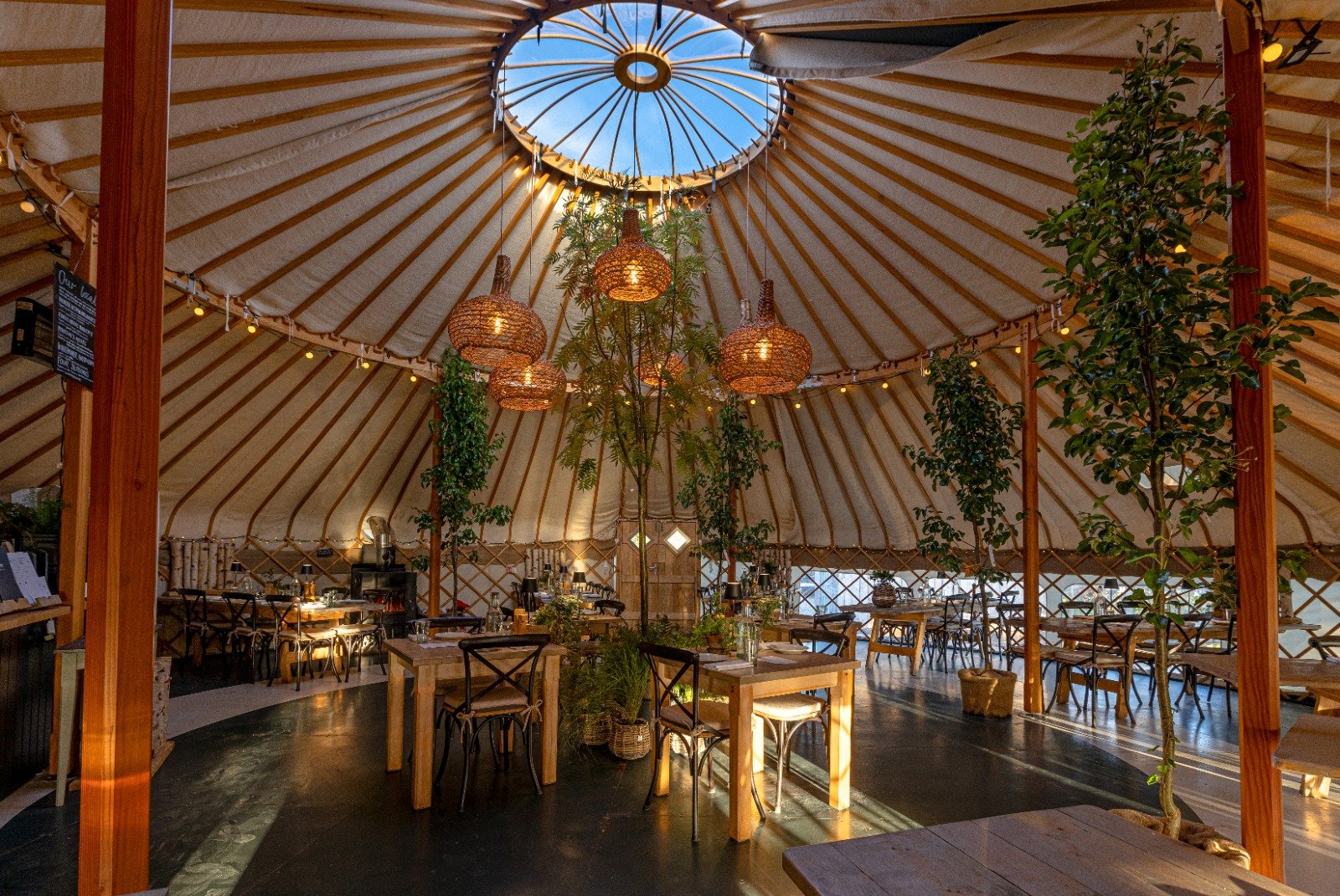 Inside our Yurt Restaurant, decorated with hanging wicker lamps and potted plants, set up as a cozy restaurant with tables and chairs.