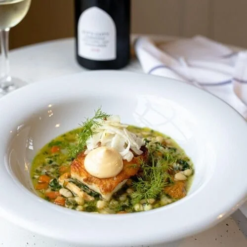 A plated fish dish with a dill sauce,  served in a white bowl. A wine bottle, wine glass, and white napkin are in the background.