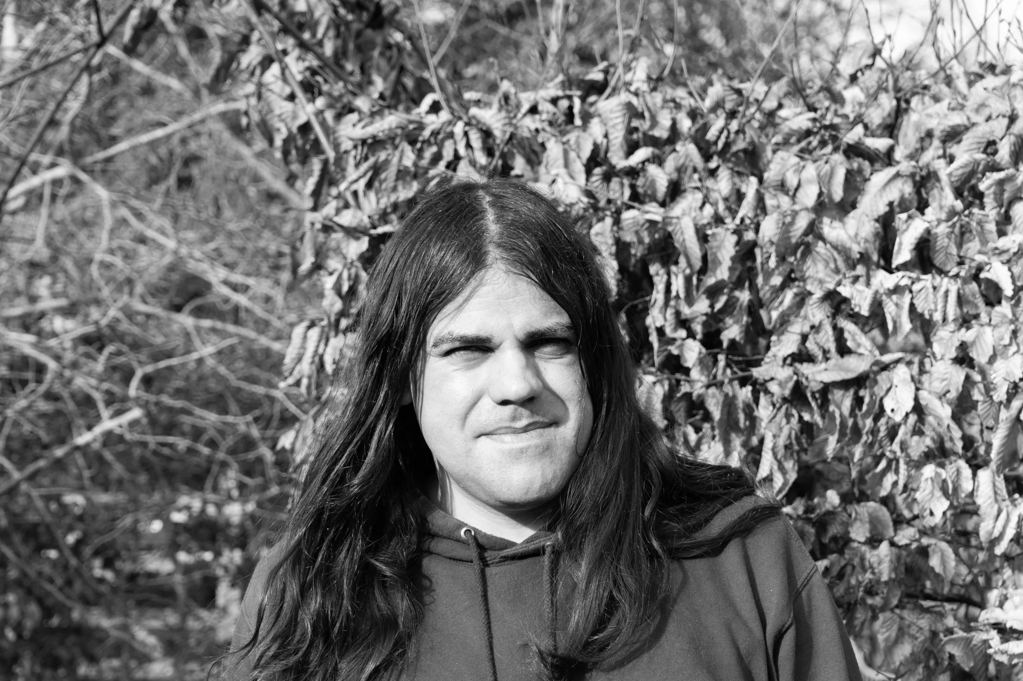 A black-and-white photo of a man with dark hair and a beard, smiling, outdoors with a blurred background of trees and foliage.