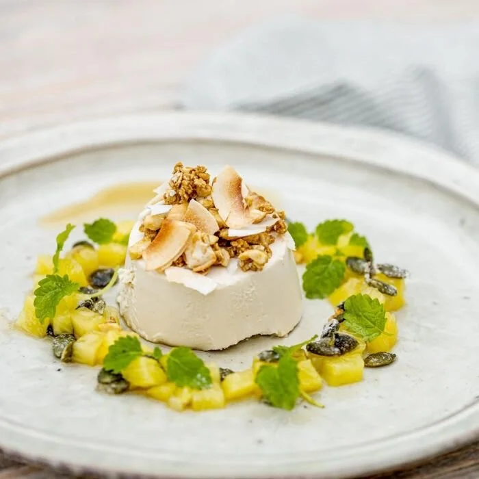 A plated dessert featuring a small white panna cotta topped with chopped nuts, surrounded by pineapple chunks, mint leaves, and black seeds on a white plate.