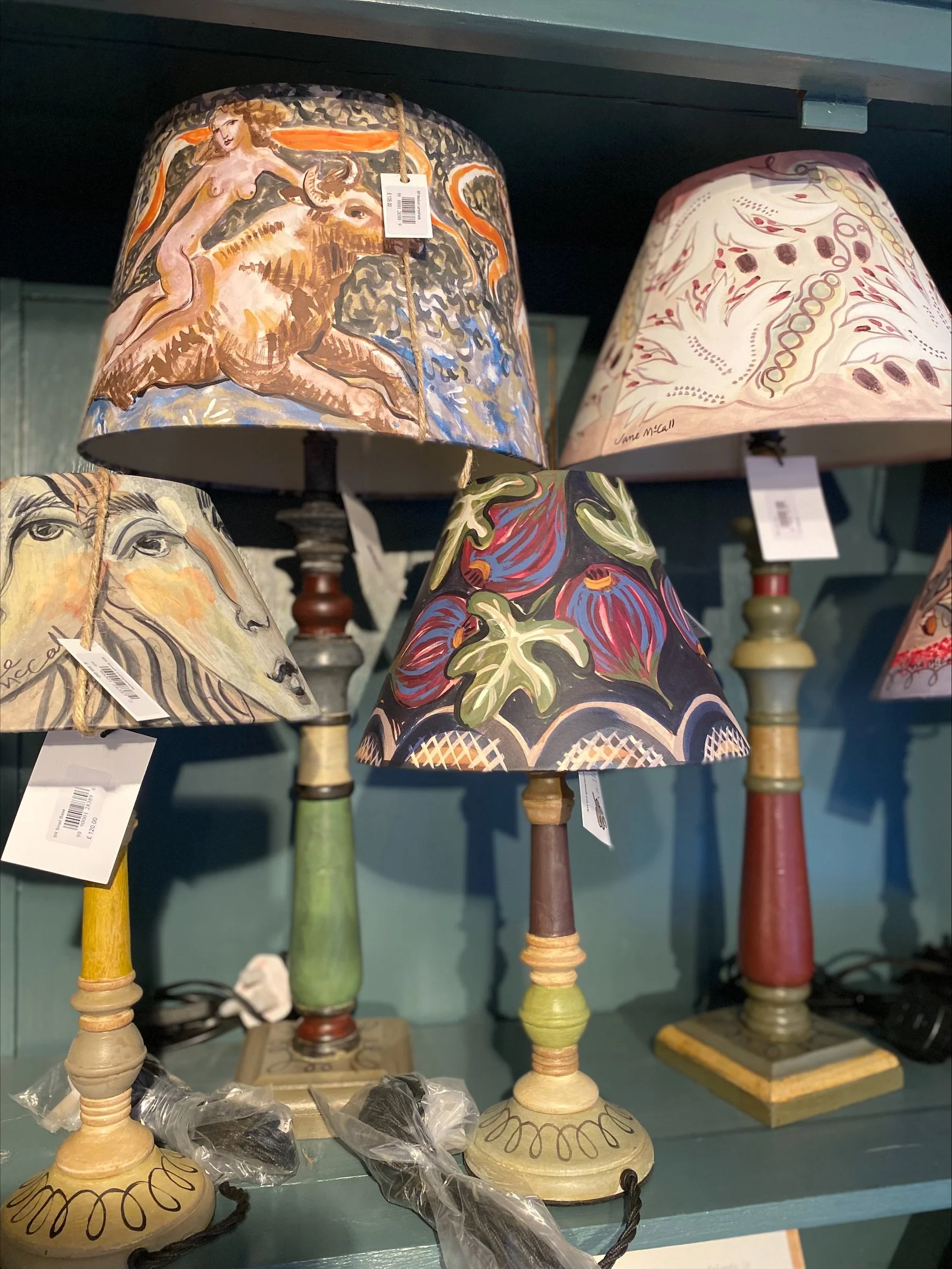 Bloomsbury revisited, Colorful table lamps with painted shades featuring various artistic and abstract designs displayed on a teal shelf.