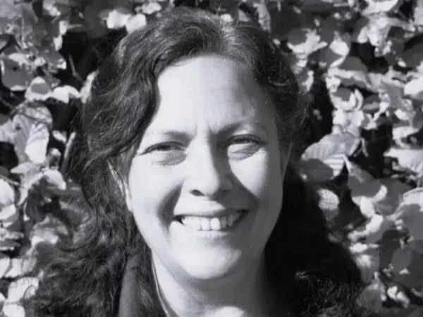 Black and white photo of a smiling woman with wavy hair, outdoors with leaves in the background.