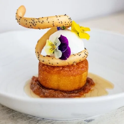 A dessert featuring a baked cake topped with a scoop of ice cream, a cookie spiral, edible flowers, and a drizzle of sauce on a white plate.