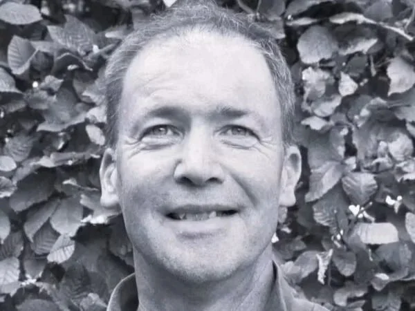 A black-and-white photo of a man smiling in front of leafy foliage.