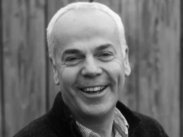 Black and white photo of a man with light-colored hair smiling, wearing a collared shirt and a dark jacket, with a wooden fence in the background.