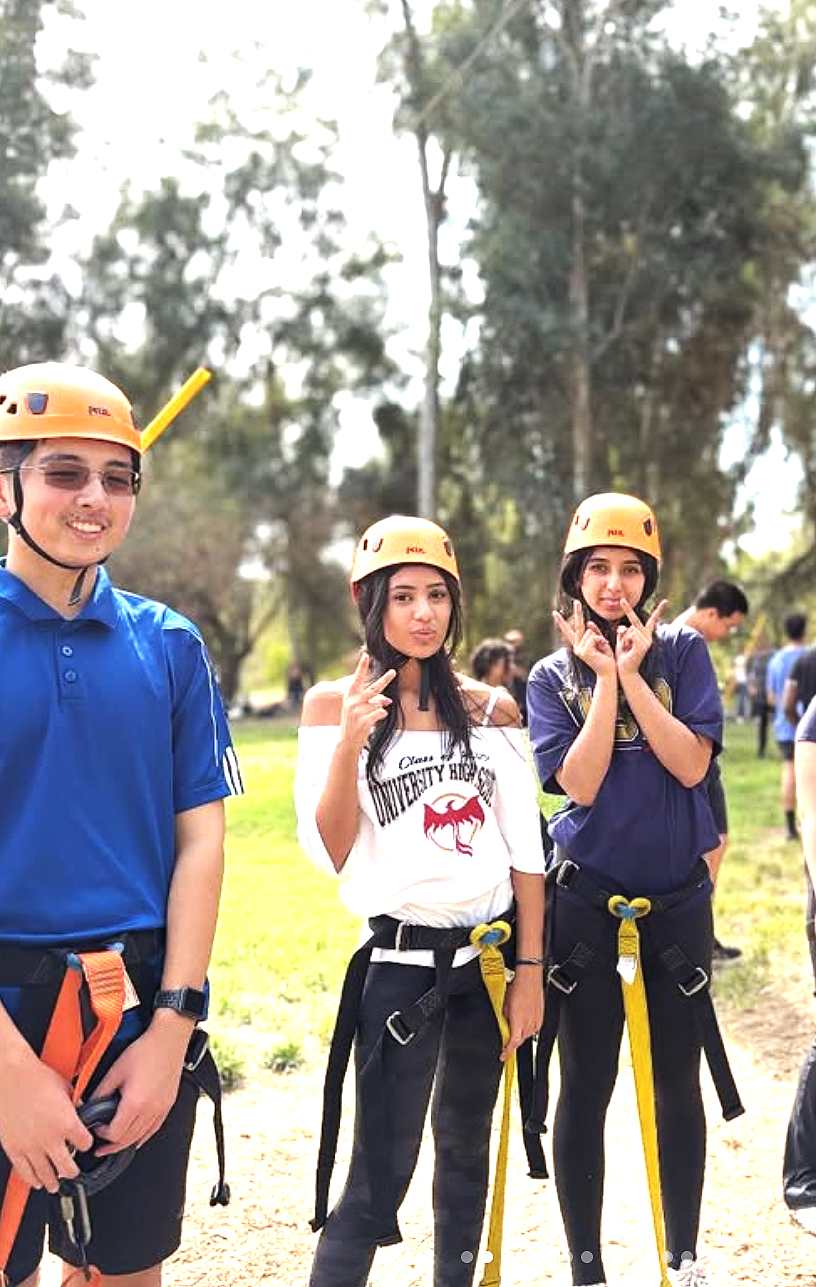 Three young adults wearing helmets and harnesses posing outdoors, with a group in the background, trees, and blue sky.