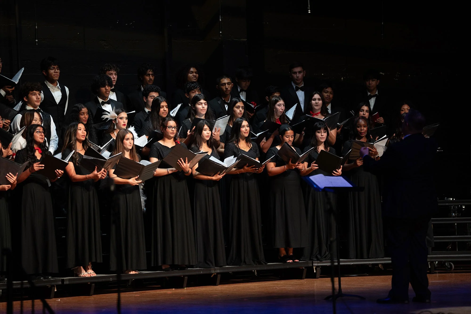 A choir of young men and women dressed in black performing on stage with a conductor in front.