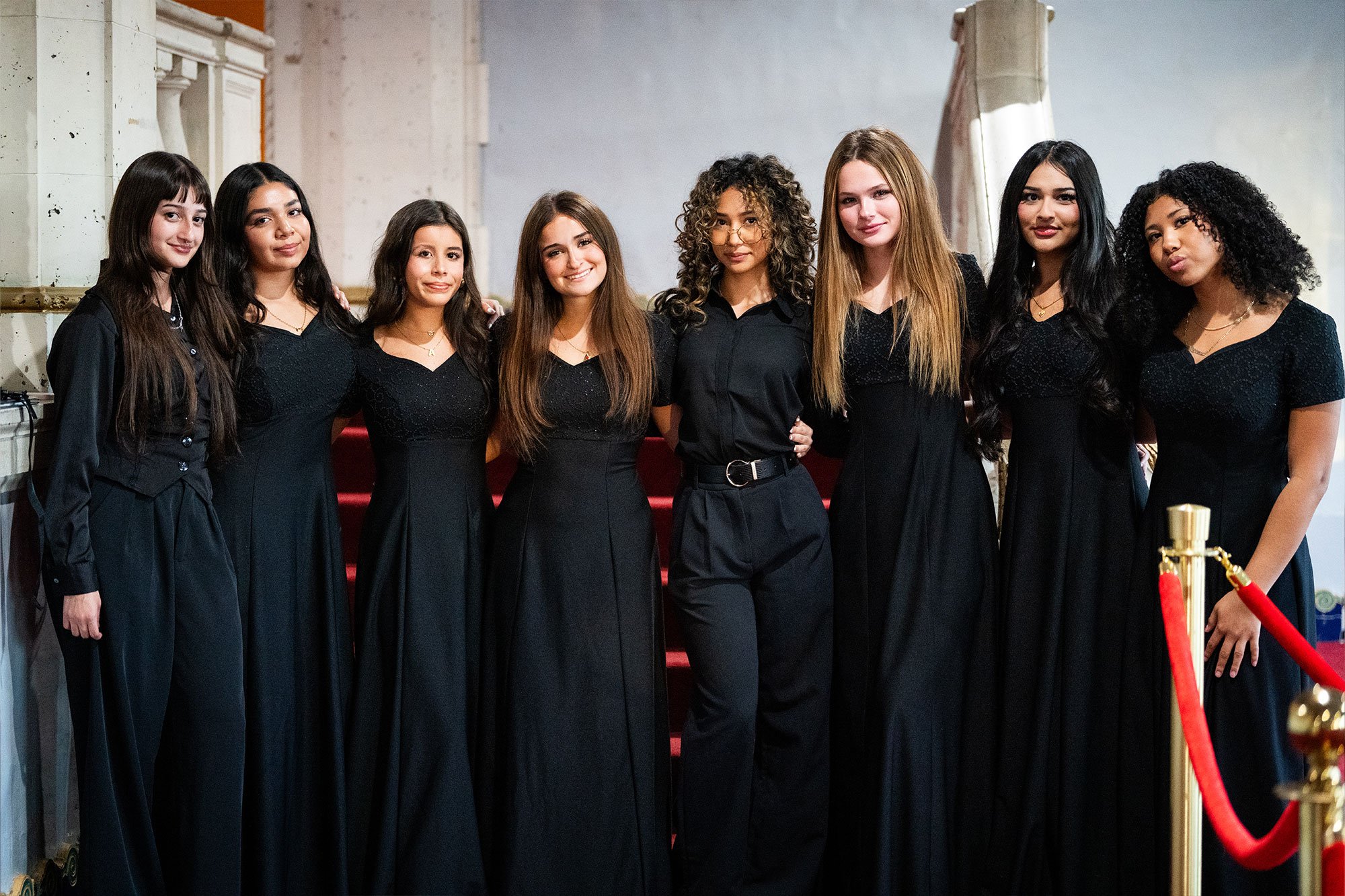 Eight women in black dresses and pants standing together in front of a staircase, posing for a group photo at an indoor event.