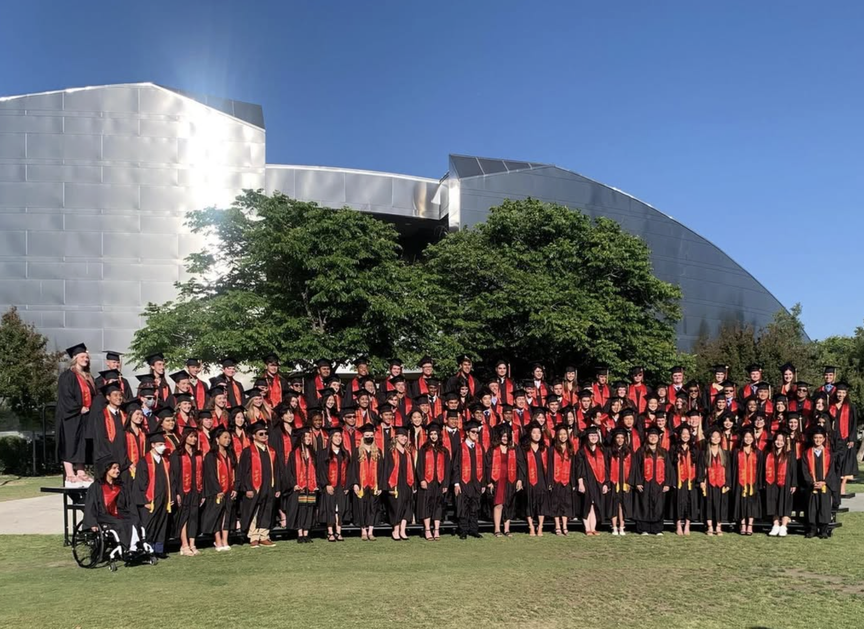 Group of graduation students in caps and gowns posing outdoors on a grassy area with modern building and trees in the background.