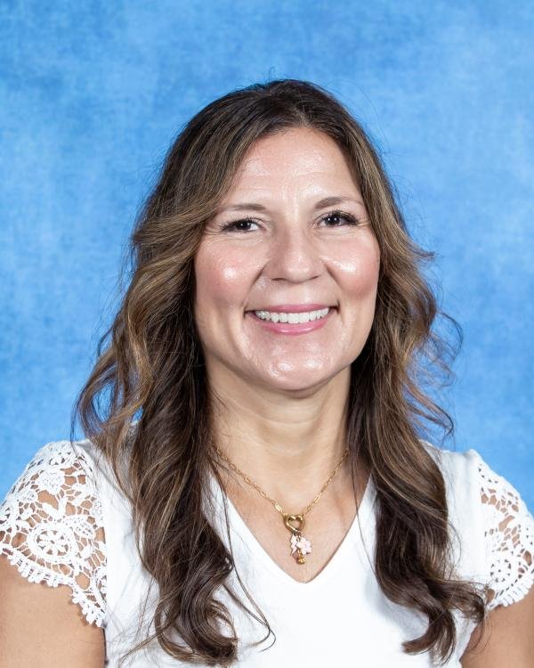 Smiling woman with wavy brown hair wearing a white lace-sleeved top and a gold necklace against a blue background.