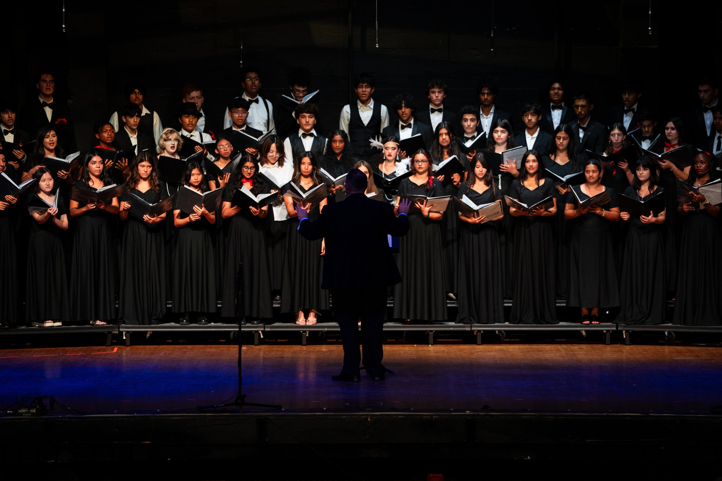 A choir of young men and women in formal black attire performing on stage, led by a conductor dressed in black, with a dark background and stage lighting.