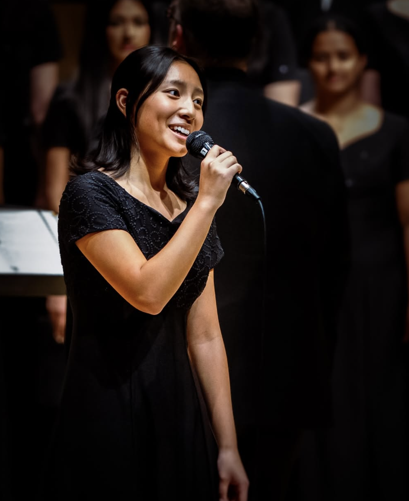 A young woman singing into a microphone on stage, wearing a black dress, with a choir behind her.