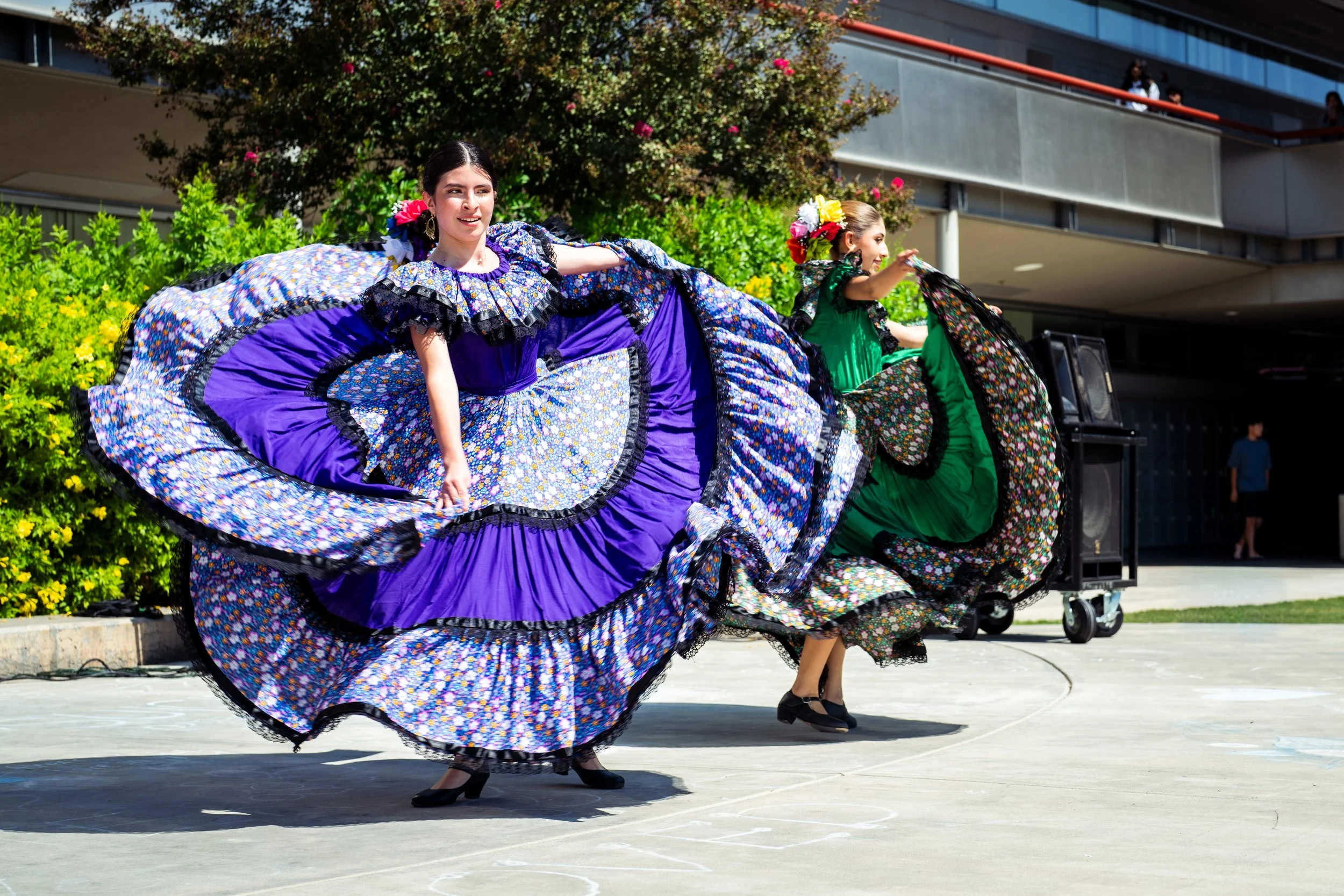 Two women and a girl dancing outdoors in traditional Mexican dresses, one in purple and the other in green, with colorful flowers in their hair, during a cultural performance.