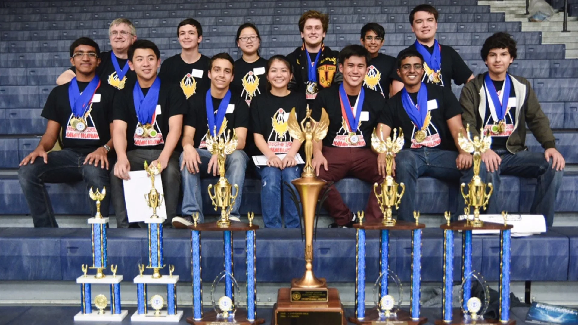Group of young people and adults sitting on blue bleachers, wearing medals and holding trophies, celebrating a victory.