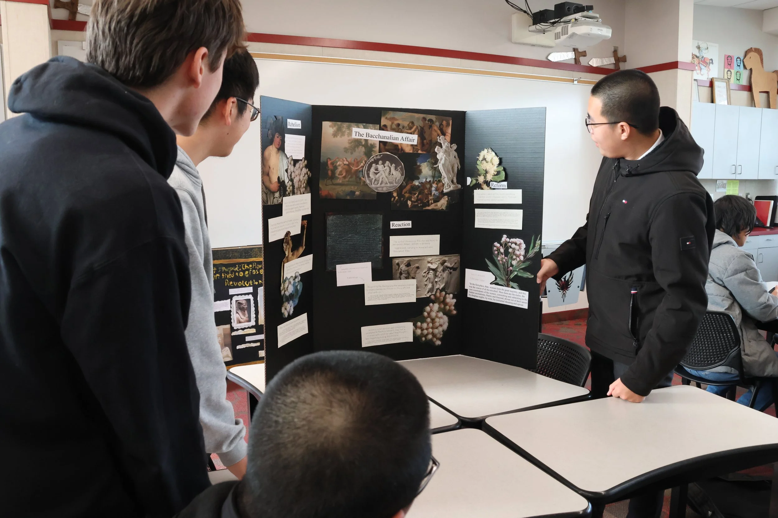 Students presenting a science project about the Bacchanalian Affair with images and texts on a trifold display board.