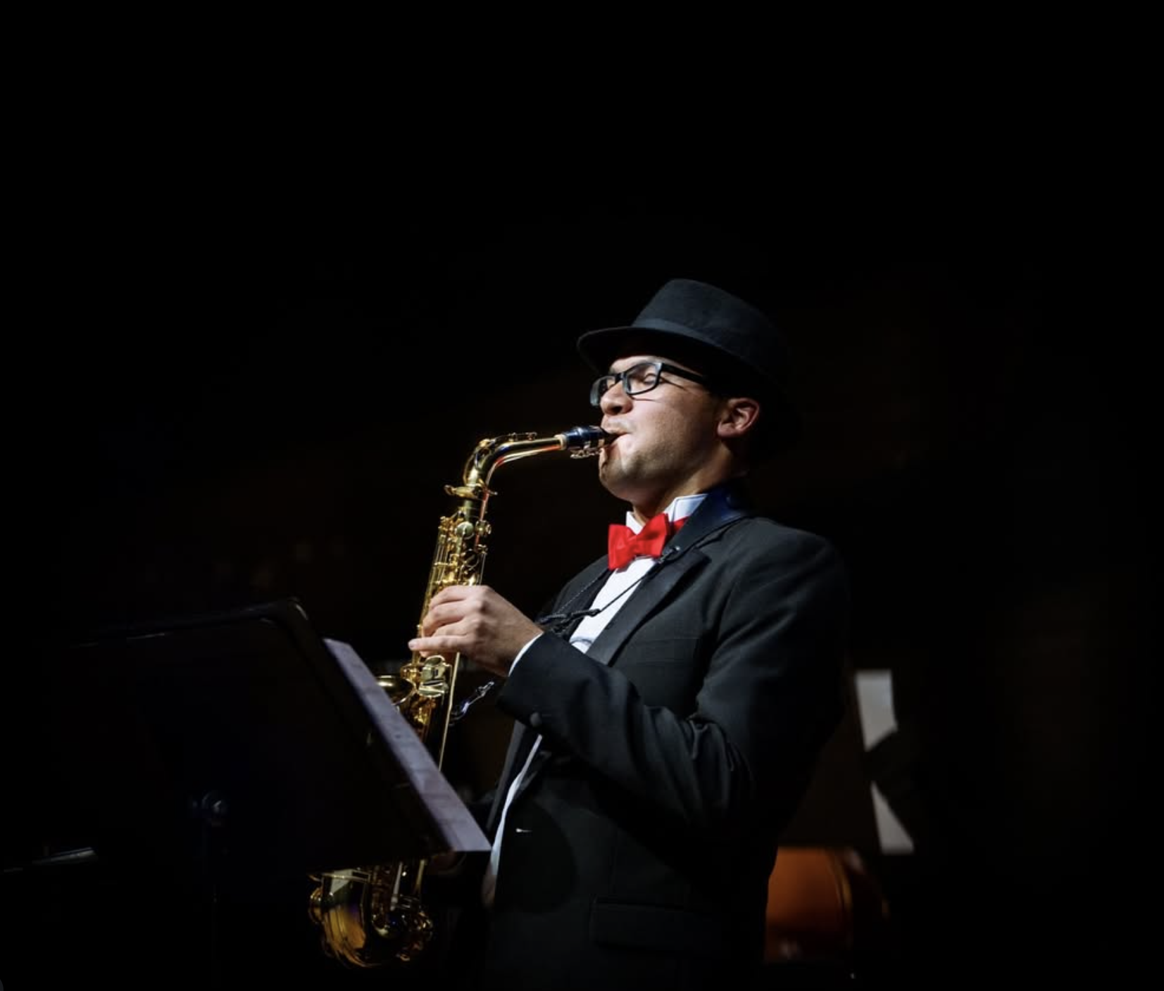 A man in a black suit, red bow tie, and black fedora playing a saxophone on stage.